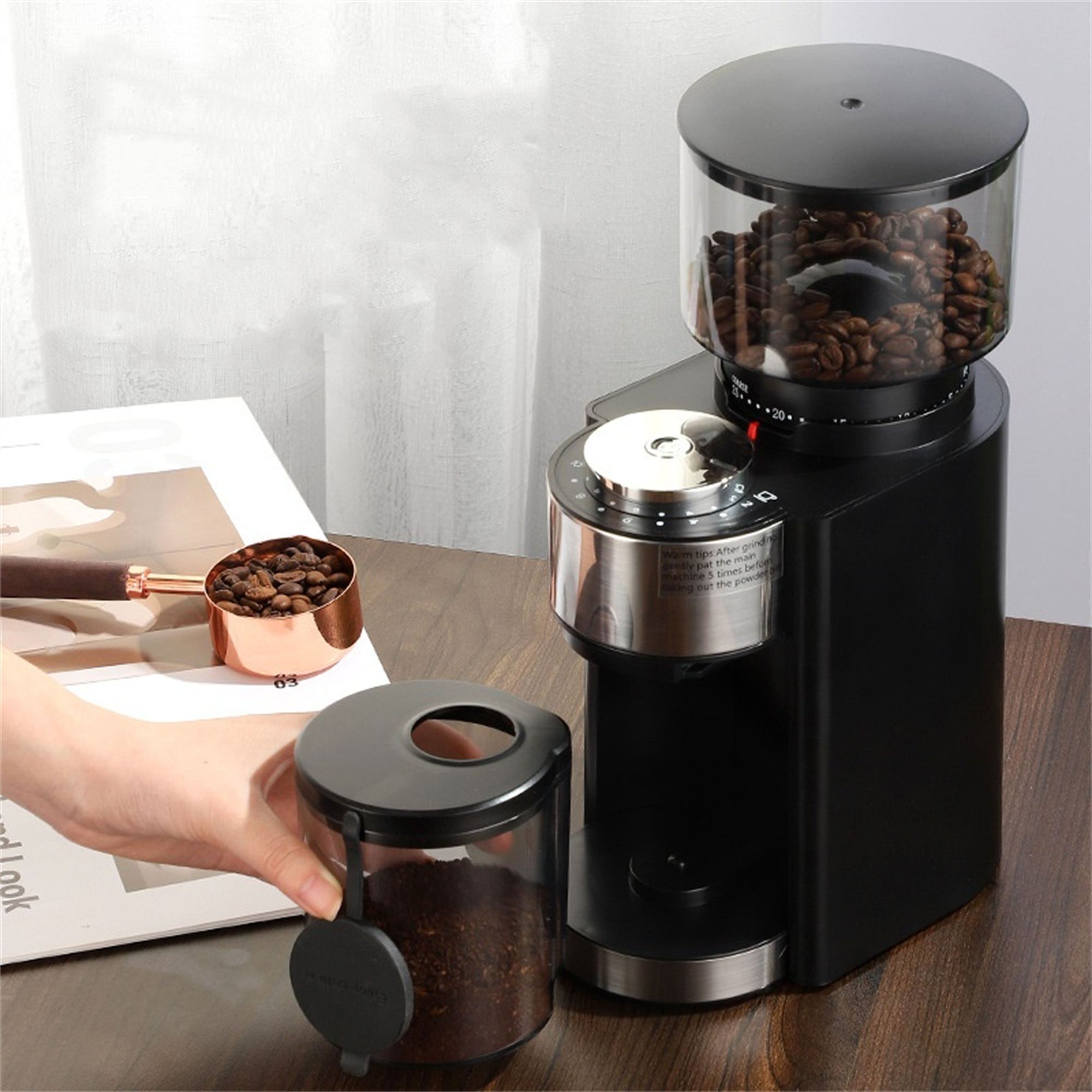 Schwarze Kaffeemühle mit transparentem Bohnenbehälter, ein Glas mit Kaffeepulver und ein Löffel mit Bohnen.