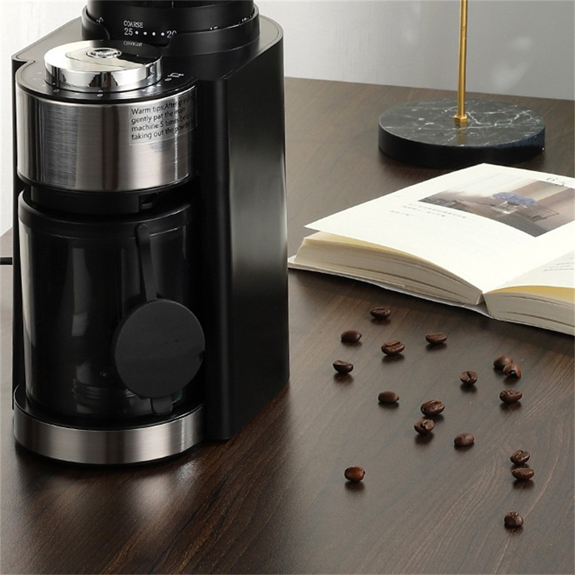 Eine schwarze Kaffeemühle mit Kaffeebohnen auf einem Holztisch und ein offenes Buch.