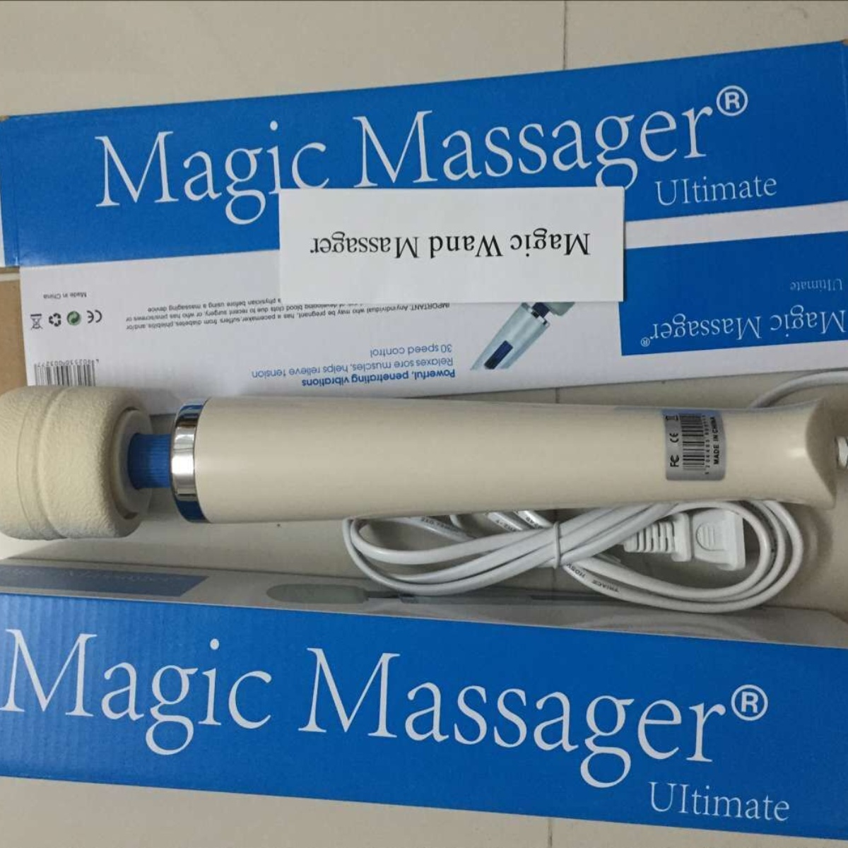 Ein weißer Massage-Zauberstab mit weichem Kopf und blauem Sockel. Er befindet sich in einer blauen Box mit Text und Etikett.