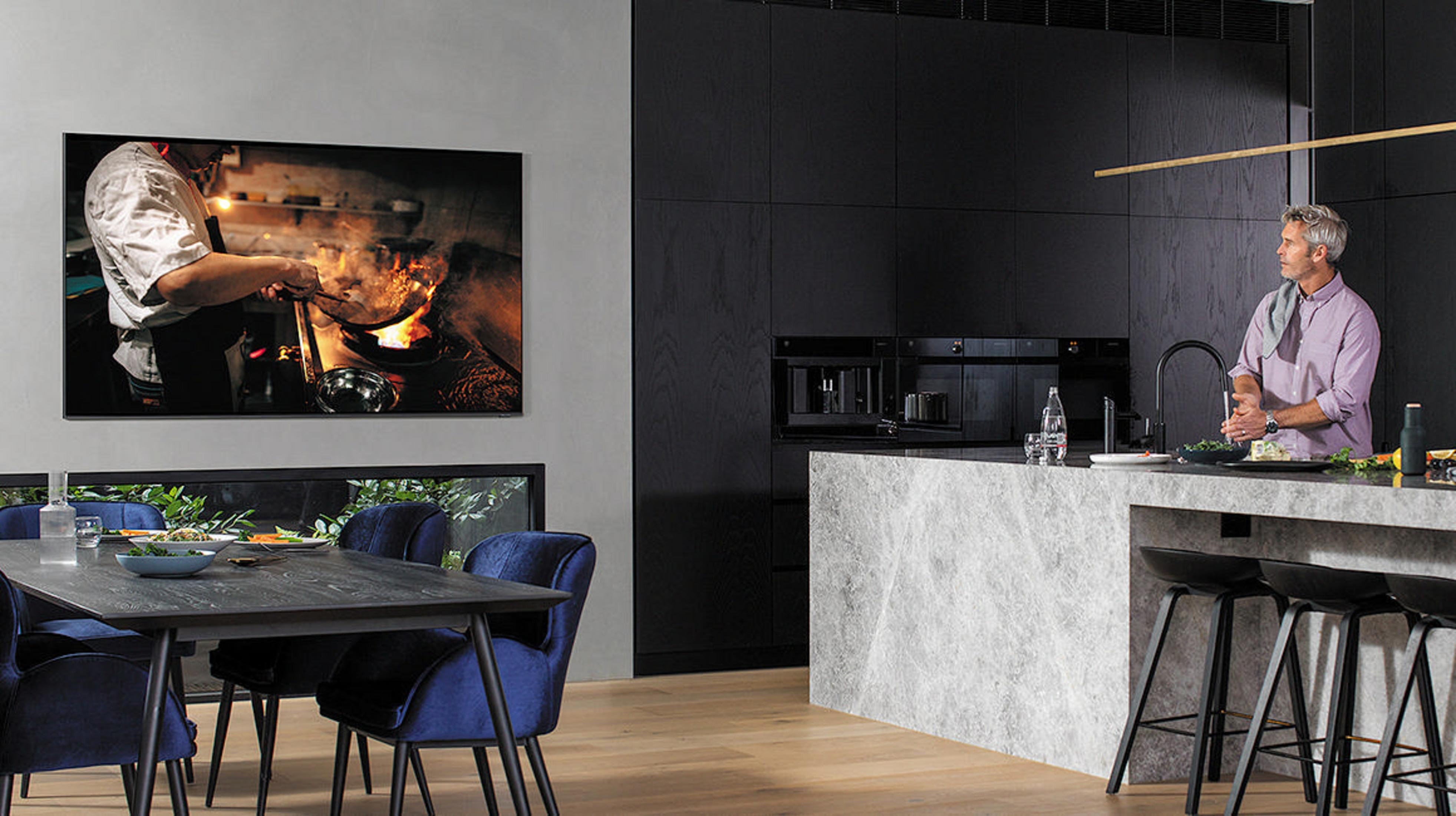 Ein Mann in einer Küche schaut fern. Der Fernseher zeigt einen Koch beim Kochen. Schwarze Schränke und Theke.
