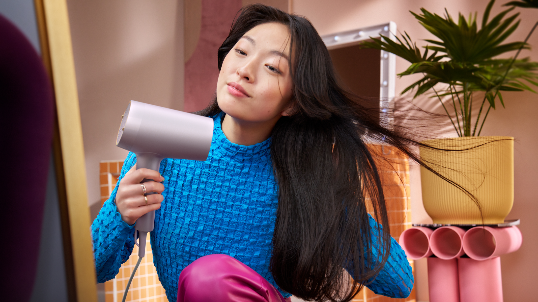 Kobieta przegląda się w lustrze, susząc włosy suszarką. Ma na sobie niebieską górę i różowe spodnie.