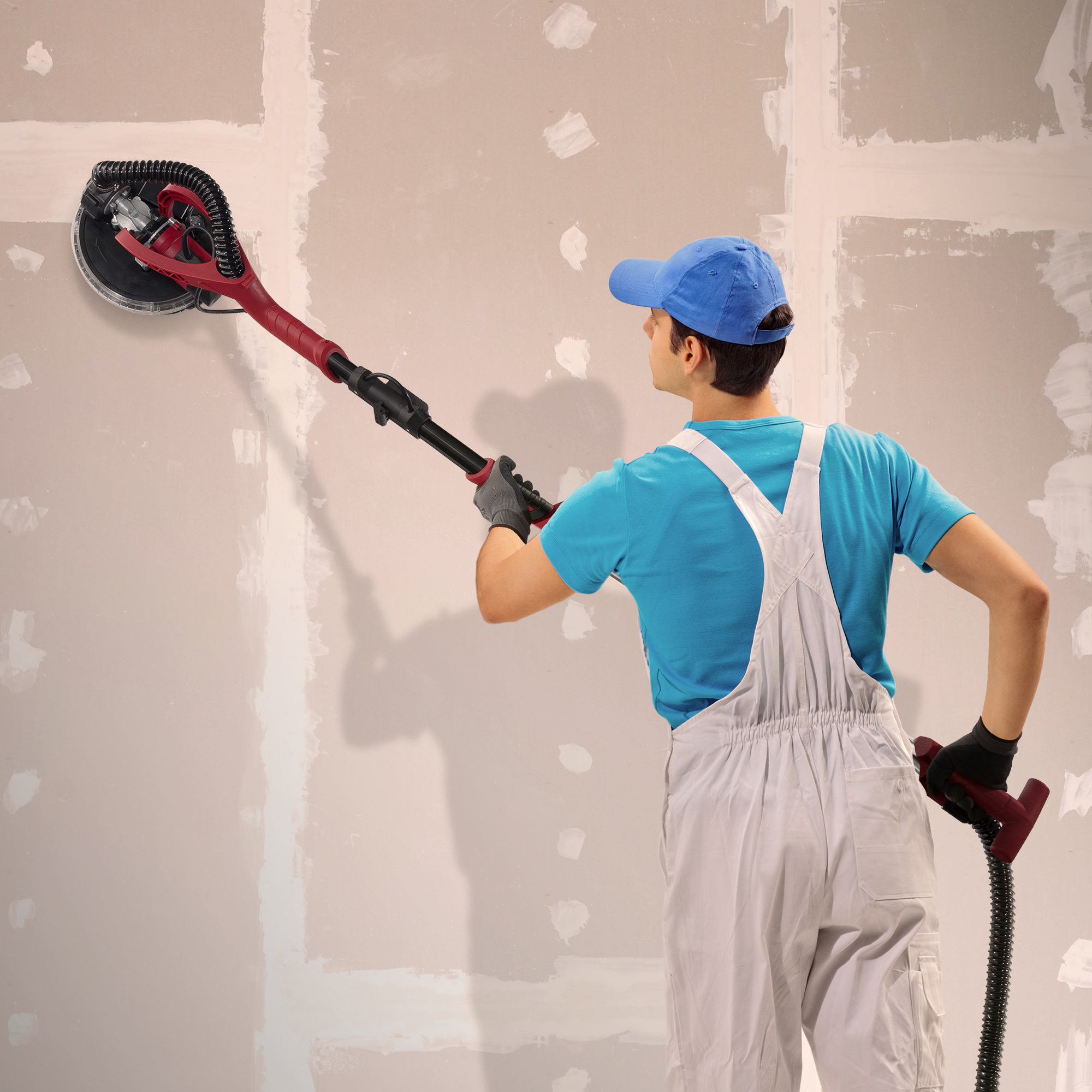 Person schleift eine Wand mit einem Trockenbauschleifer. Der Arbeiter trägt eine blaue Mütze, einen Overall und Handschuhe.