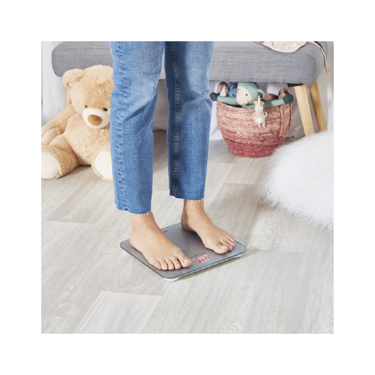 Osoba stoi na wadze cyfrowej na podłodze o wyglądzie drewna, z misiem, ławką i dekoracjami w tle.