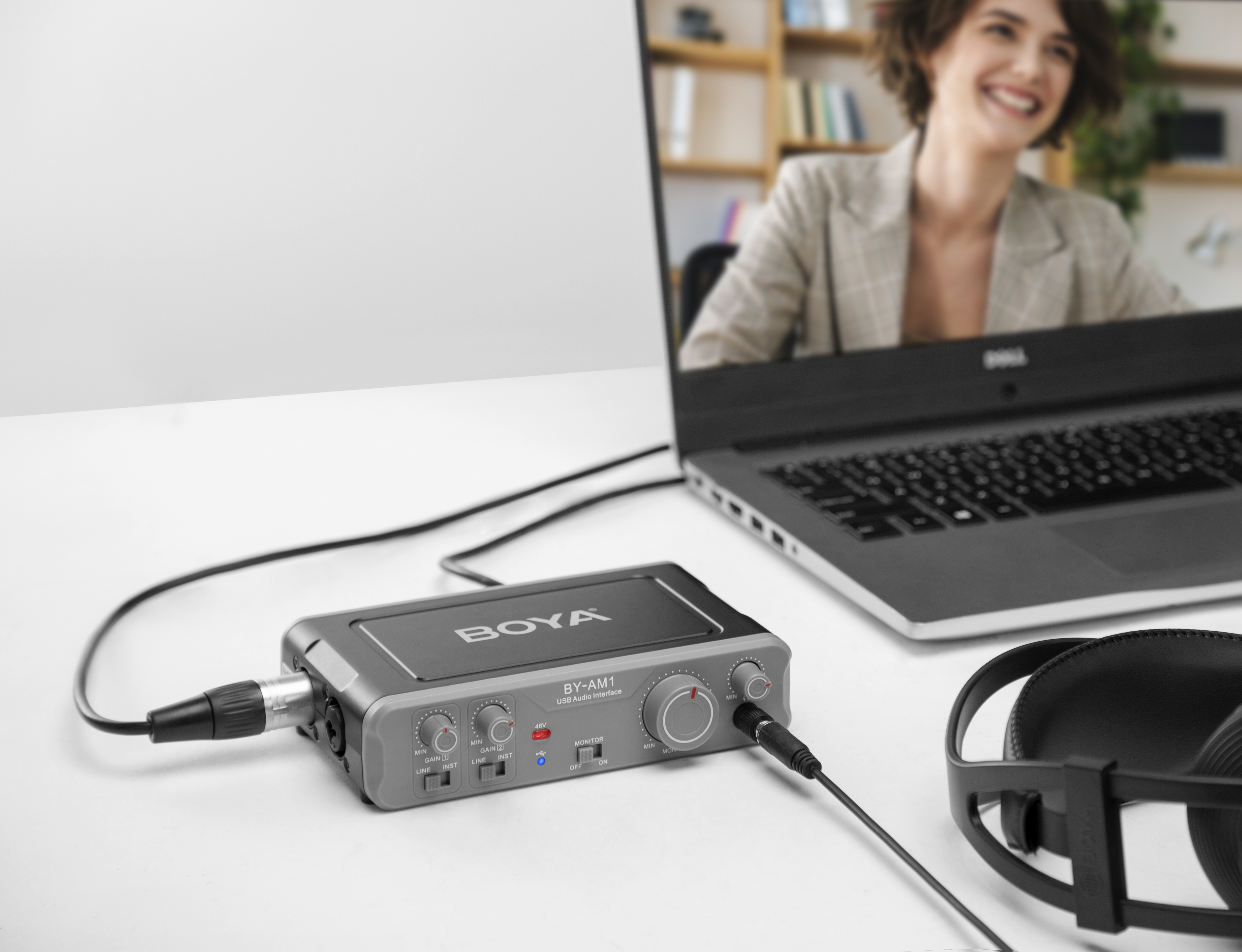 Una interfaz de audio plateada, laptop y auriculares sobre un escritorio blanco. Una mujer está en la pantalla de la laptop.