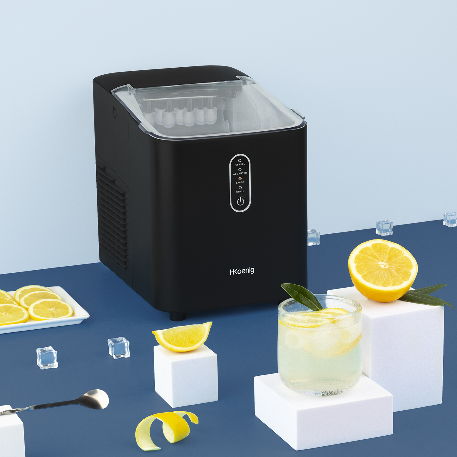 Macchina per il ghiaccio nera. Fette di limone e un bicchiere con una bevanda al limone. Cubetti bianchi su una superficie blu.