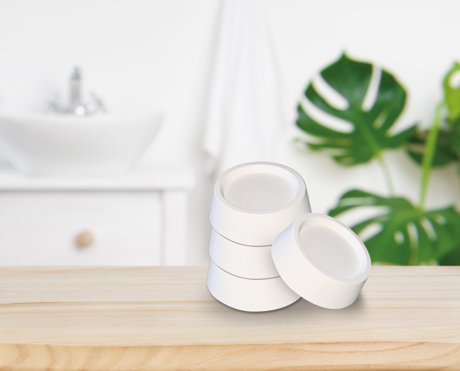 Una pila de cuatro recipientes blancos y redondos sobre una mesa de madera. Un lavabo y una planta están en el fondo.