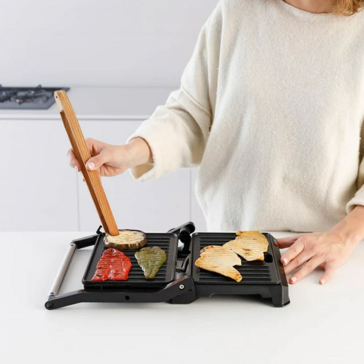 Osoba grilluje warzywa na czarnym grillu. Osoba używa drewnianych szczypiec do manipulowania jedzeniem na grillu.