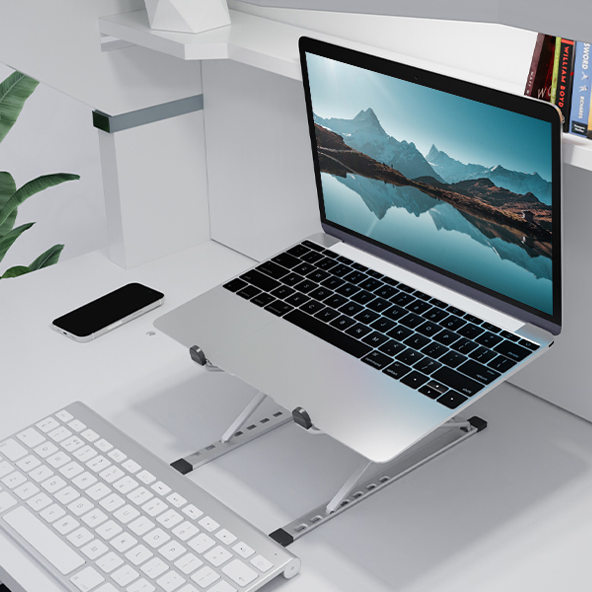 Laptop auf einem Ständer, Tastatur, Telefon auf einem weißen Schreibtisch. Pflanze und Bücher im Hintergrund.