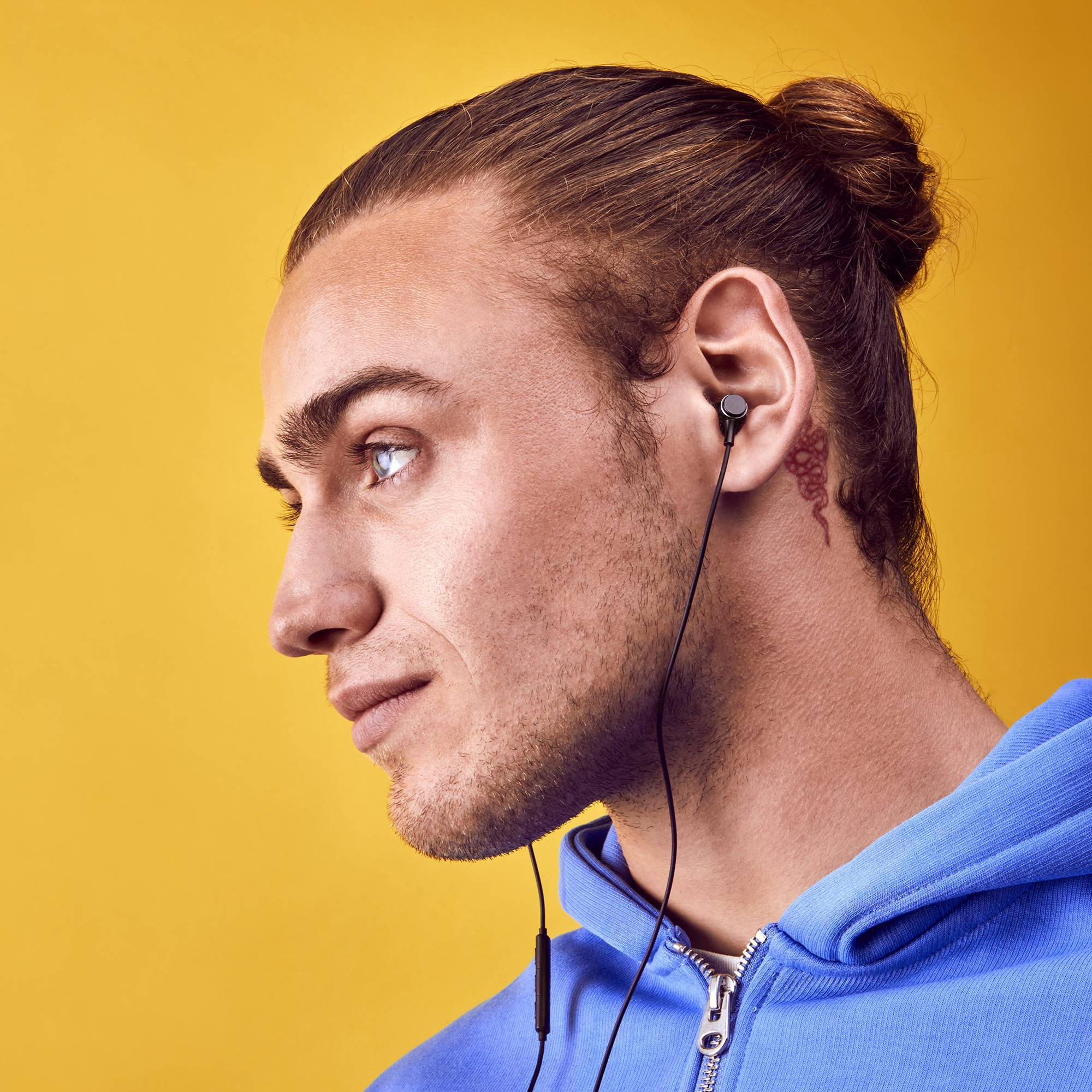 Ein Mann mit Dutt und Kopfhörern. Der Mann trägt einen blauen Kapuzenpullover, der Hintergrund ist gelb.