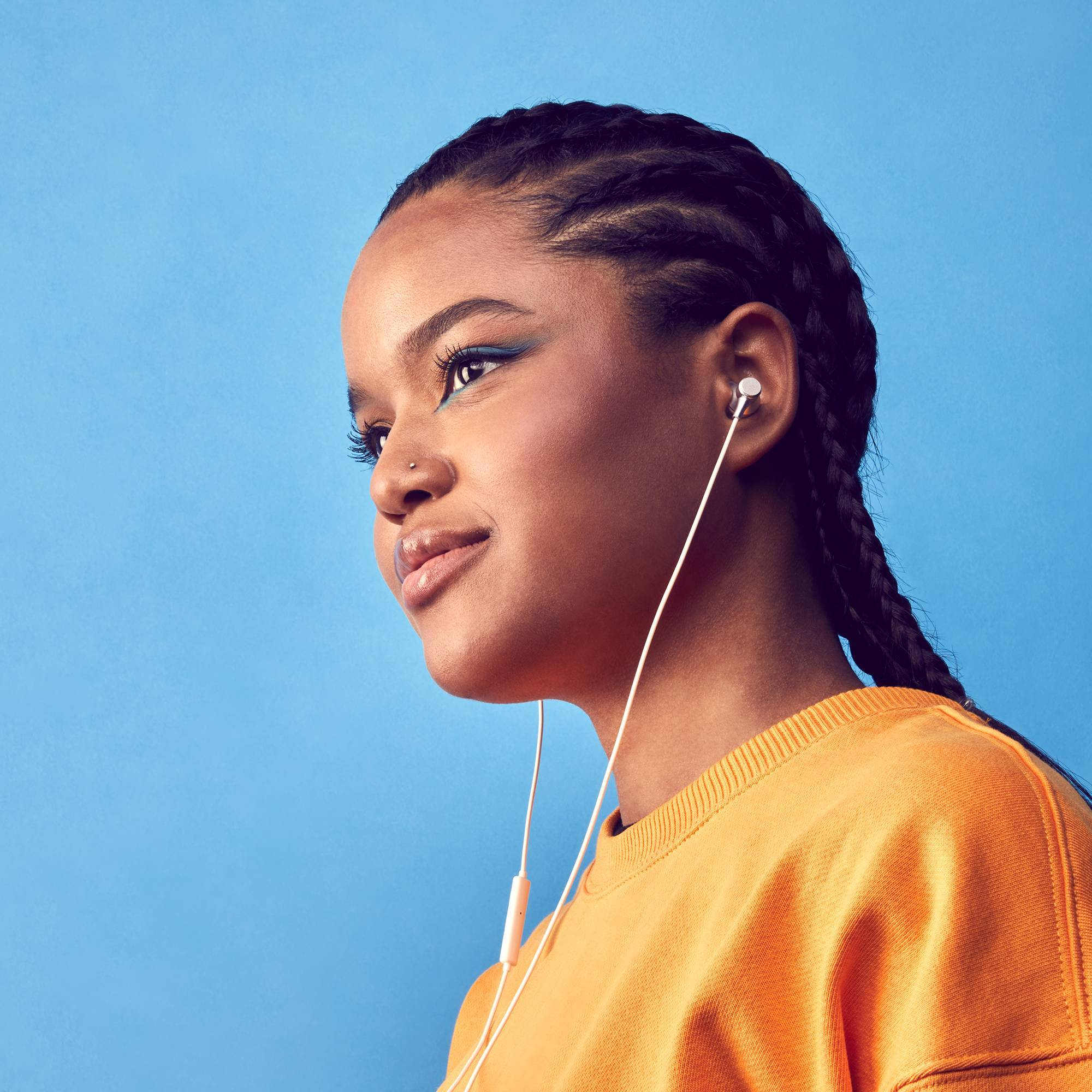 Una joven con trenzas que usa una camiseta naranja, escuchando música con auriculares blancos.