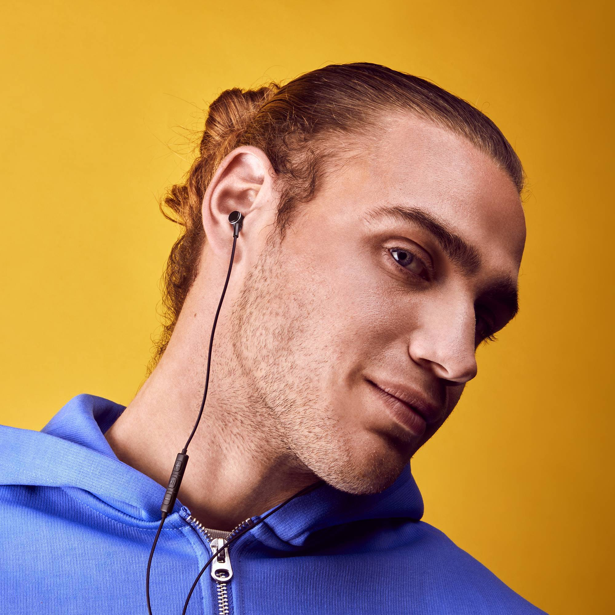 Ein junger Mann mit blauem Kapuzenpullover und Ohrhörern im Ohr, lächelnd. Gelber Hintergrund.