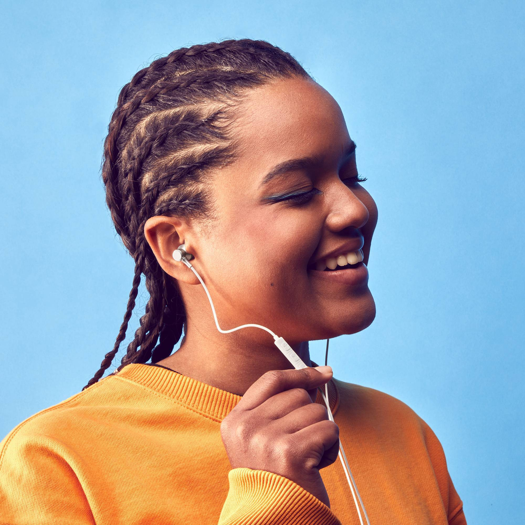 Una mujer sonriente con trenzas usa un suéter naranja, escuchando música con auriculares blancos.
