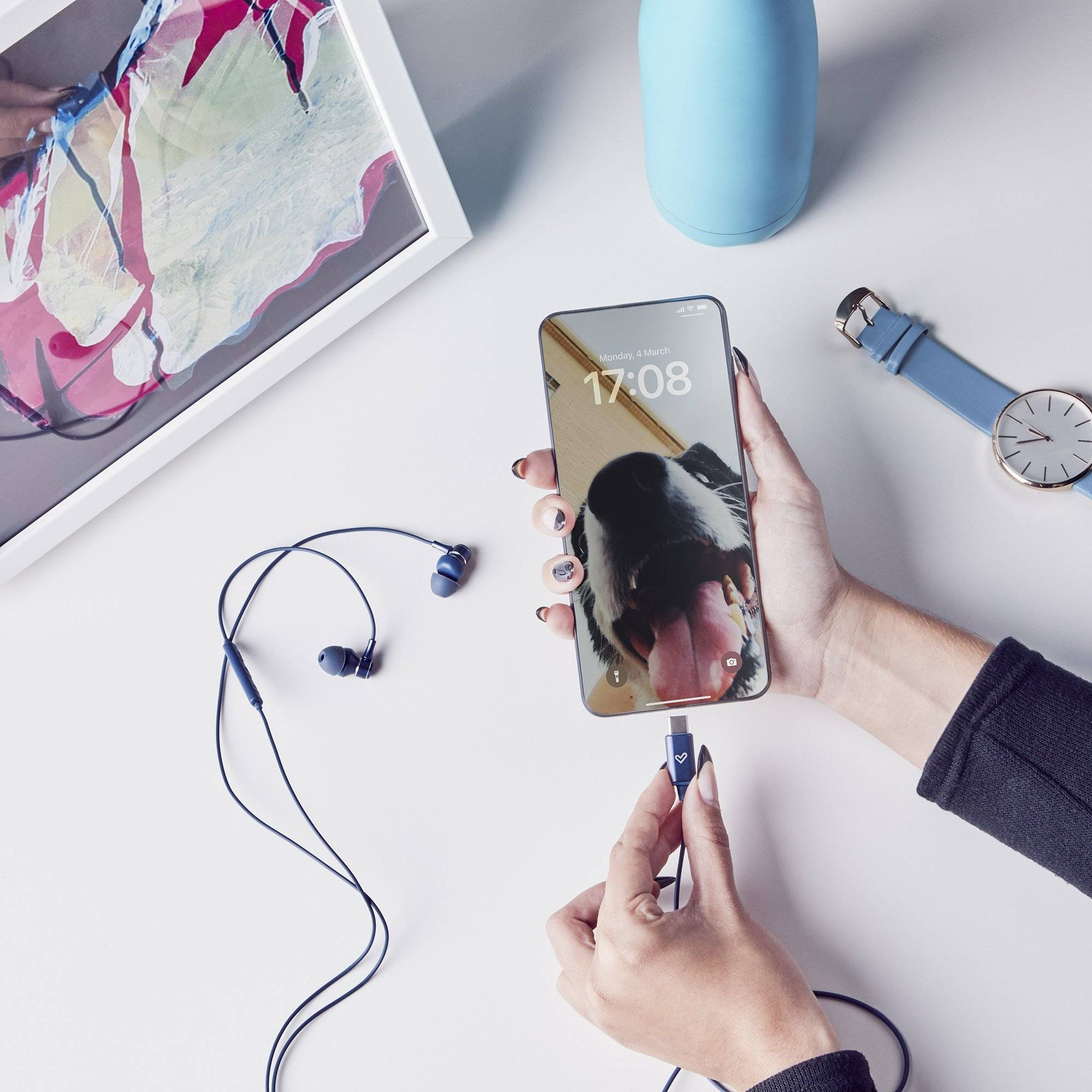 Mani che tengono un telefono con foto di cane. Auricolari e orologio su superficie bianca.