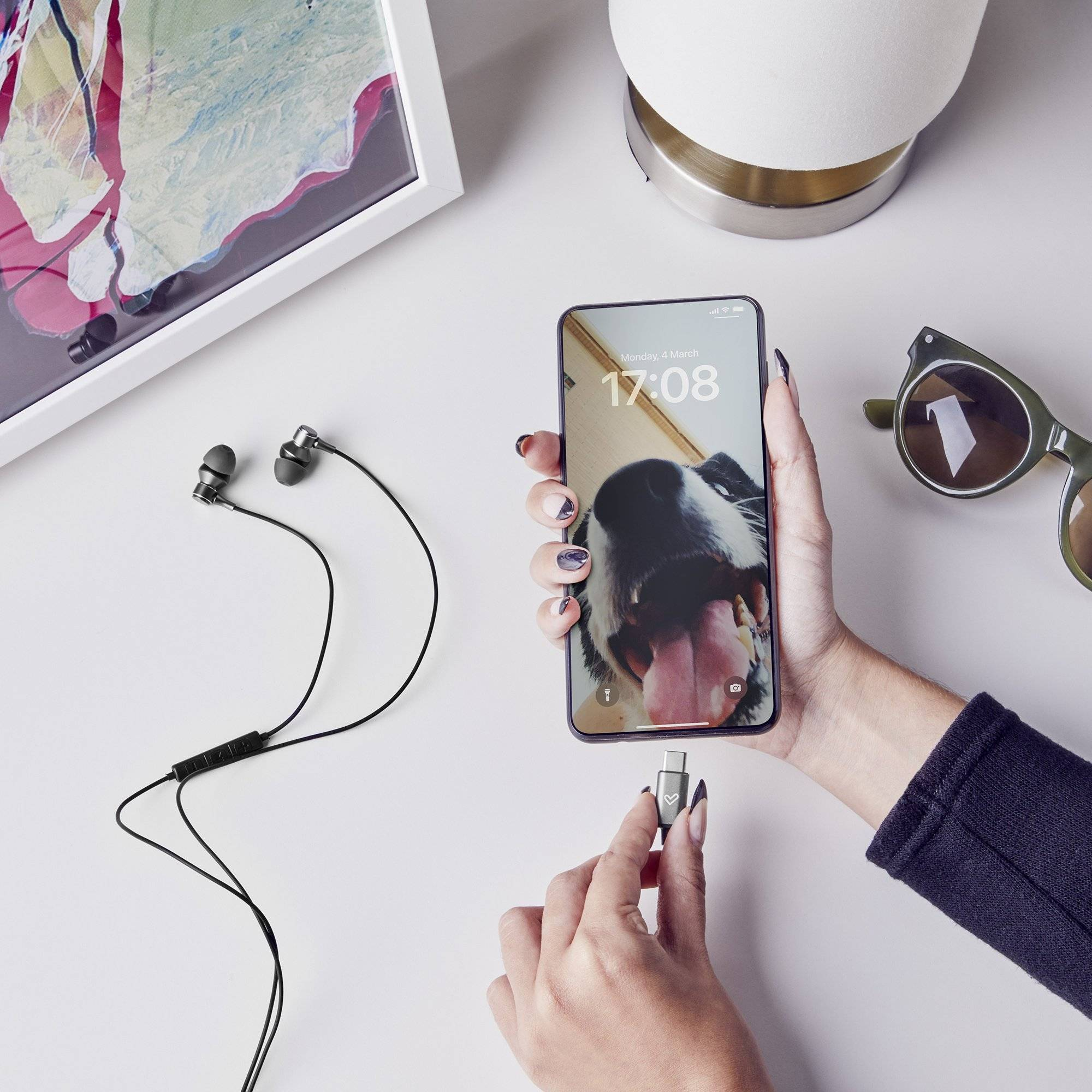 Persona sosteniendo un teléfono con imagen de perro, conectado a un cable USB-C y auriculares en superficie blanca.