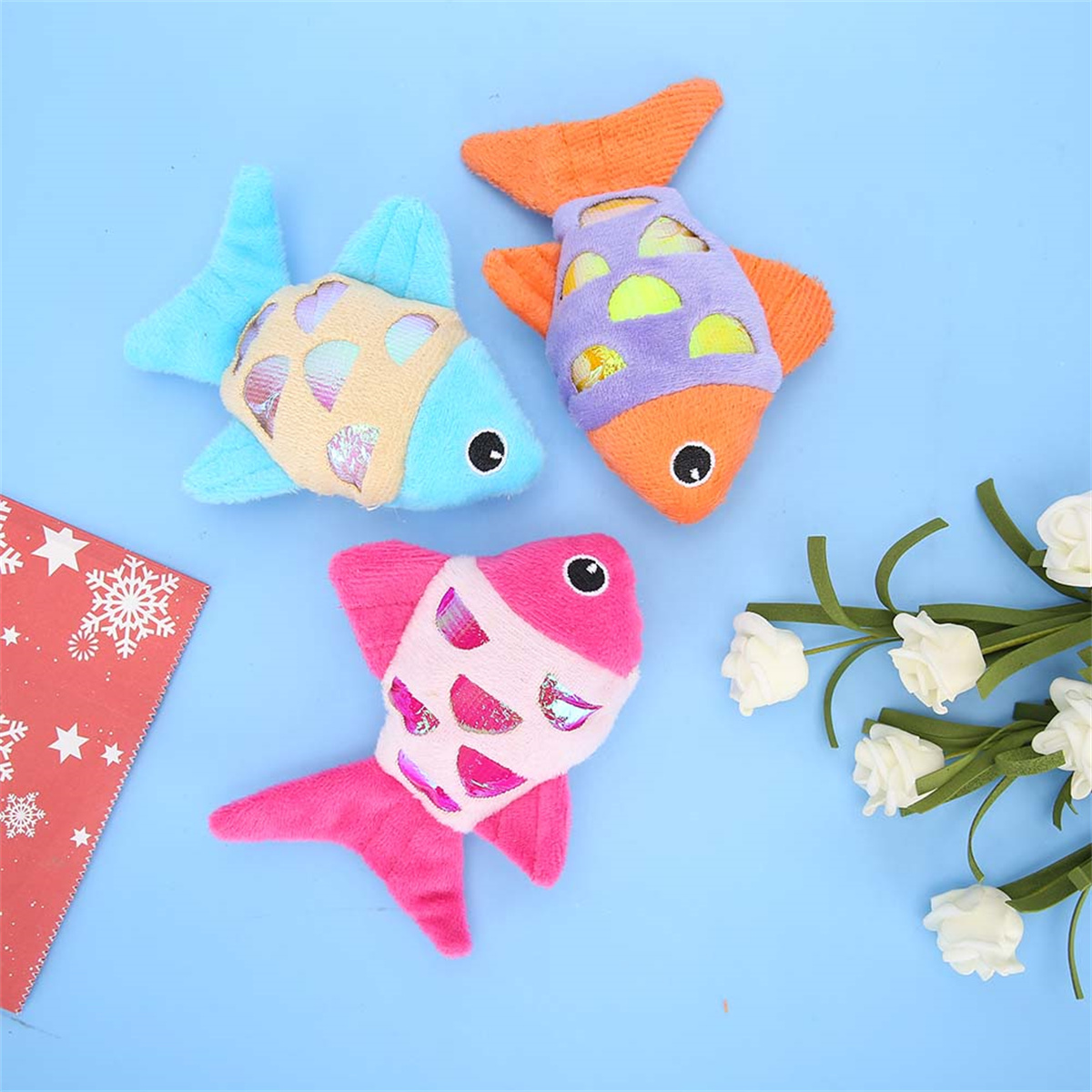 Drei bunte Fisch-Haustierspielzeuge auf einer blauen Oberfläche. Weiße Blumen und eine rote Karte sind sichtbar.