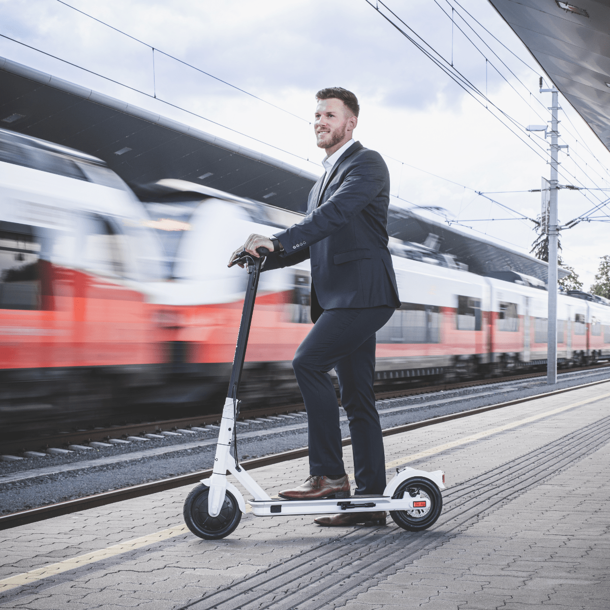 Mann auf weißem Roller am Bahnhof, Zug im Hintergrund verschwommen.
