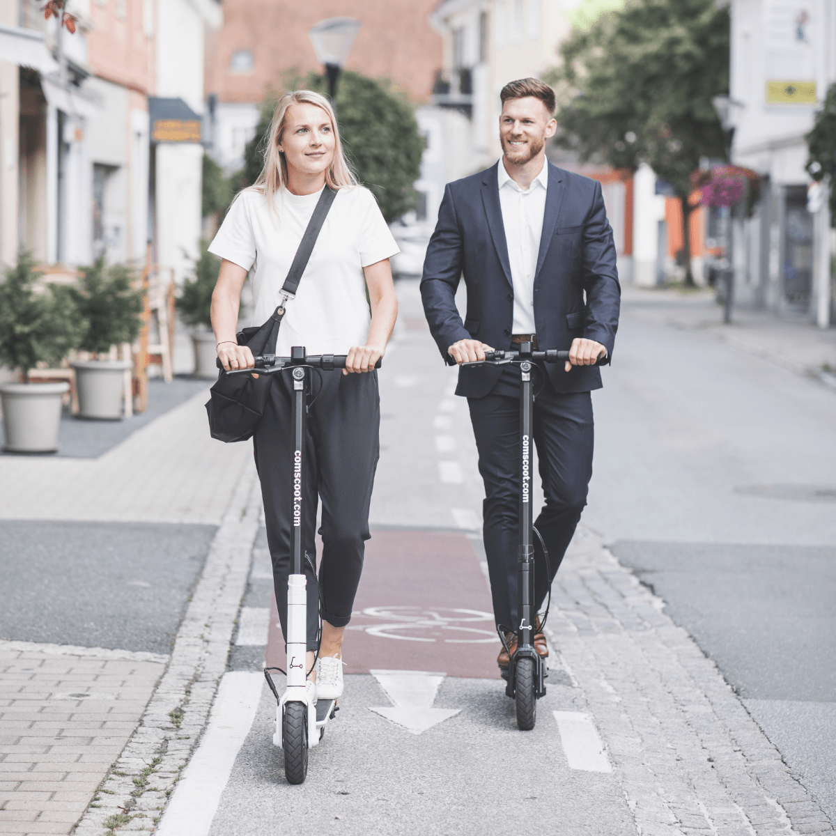 Ein Mann und eine Frau fahren mit Rollern auf einer Straße. Gebäude und Bäume säumen die Straße.