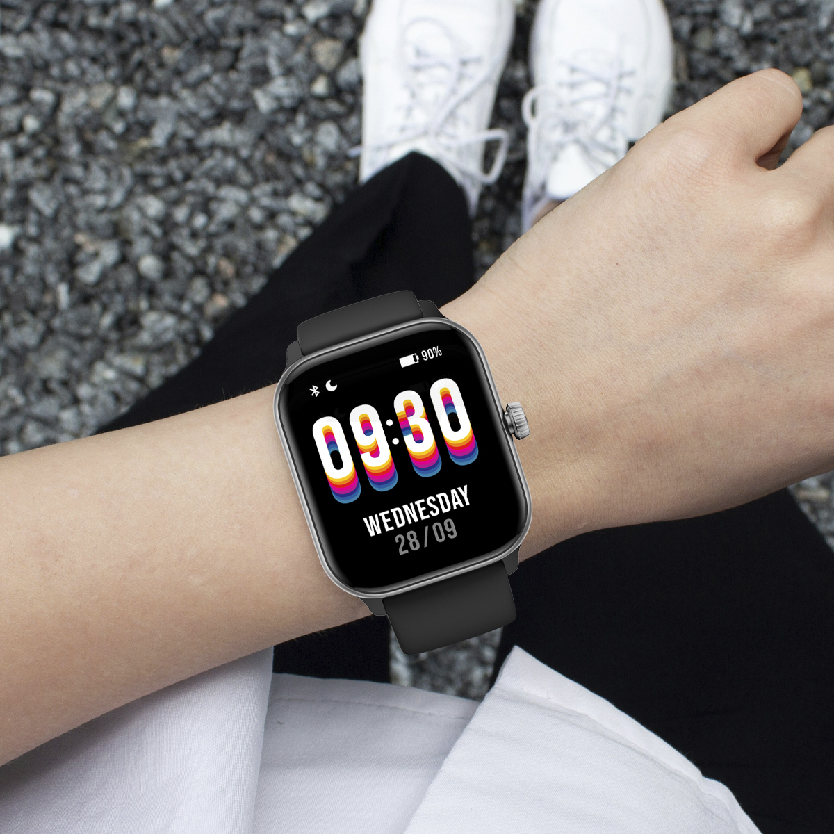 Osoba nosząca czarny smartwatch pokazujący godzinę. Ich ręce są na nogach i noszą białe buty.