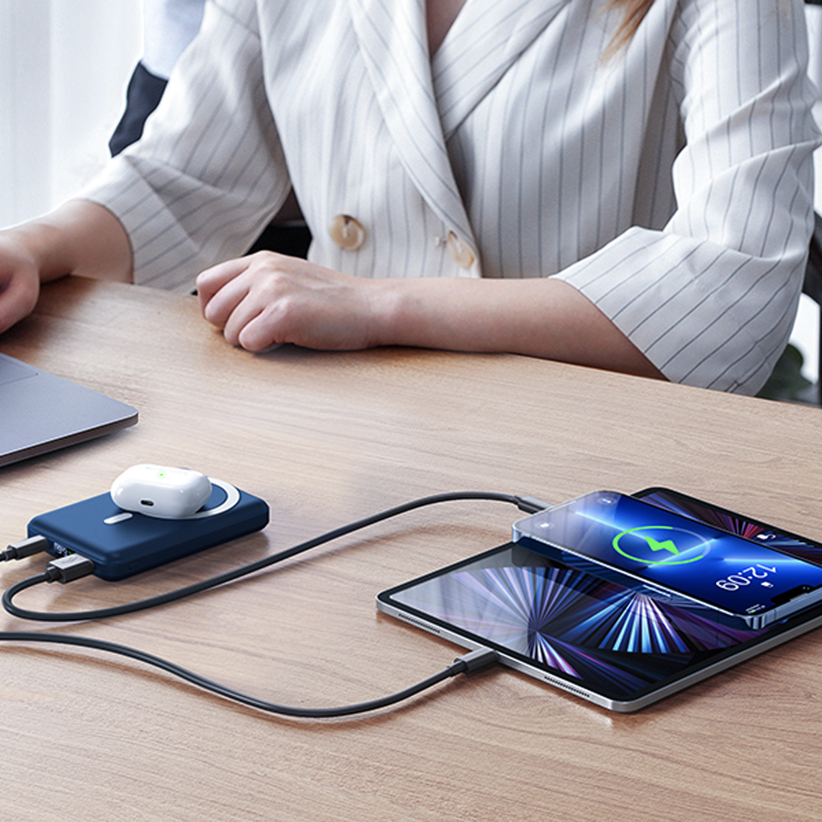 Eine Person lädt Geräte auf einem Schreibtisch auf. Ein Tablet und AirPods sind sichtbar. Eine blaue Powerbank wird verwendet.