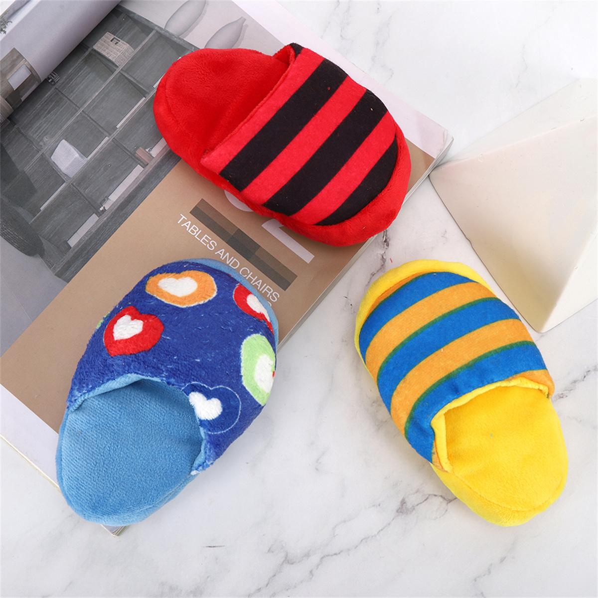 Drei bunte Hausschuhe: rot mit schwarzen Streifen, blau mit Herzen und gelb mit blauen Streifen, auf gemusterter Oberfläche.