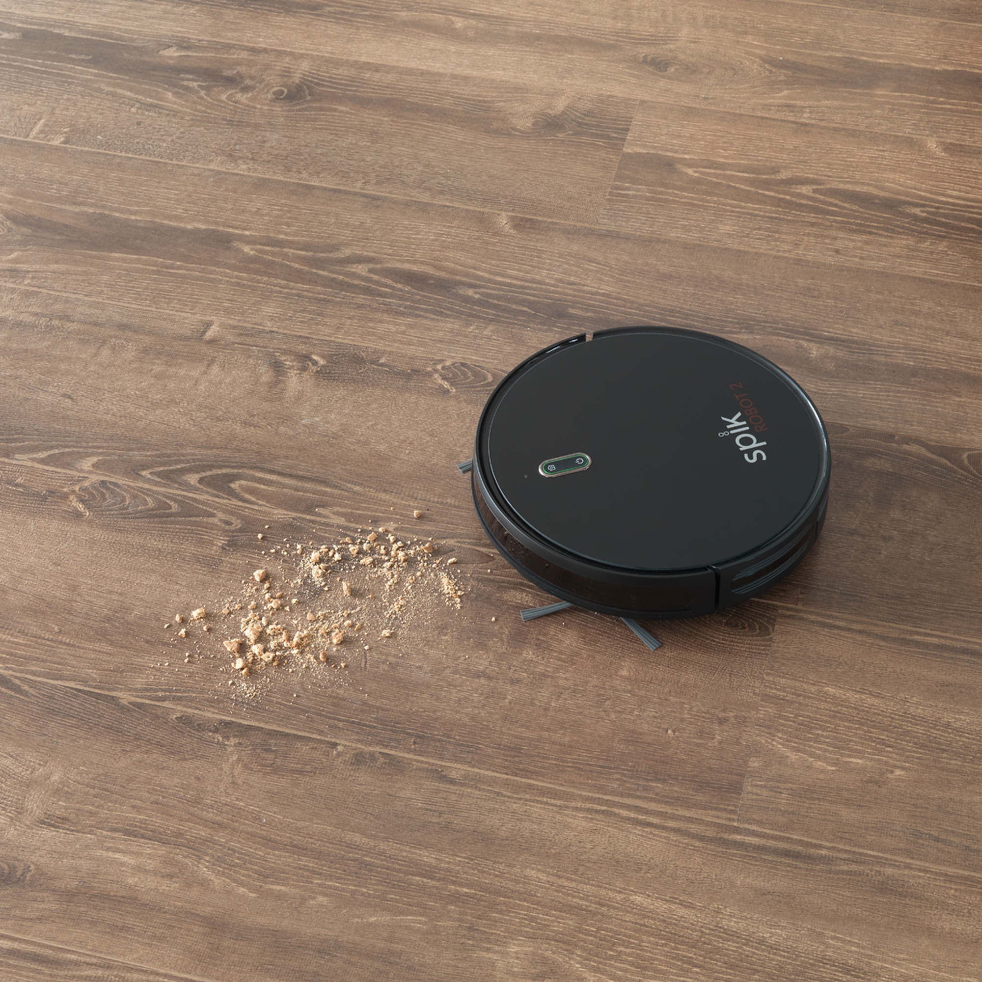 Aspiradora robot negra en suelo de madera, con restos cerca.