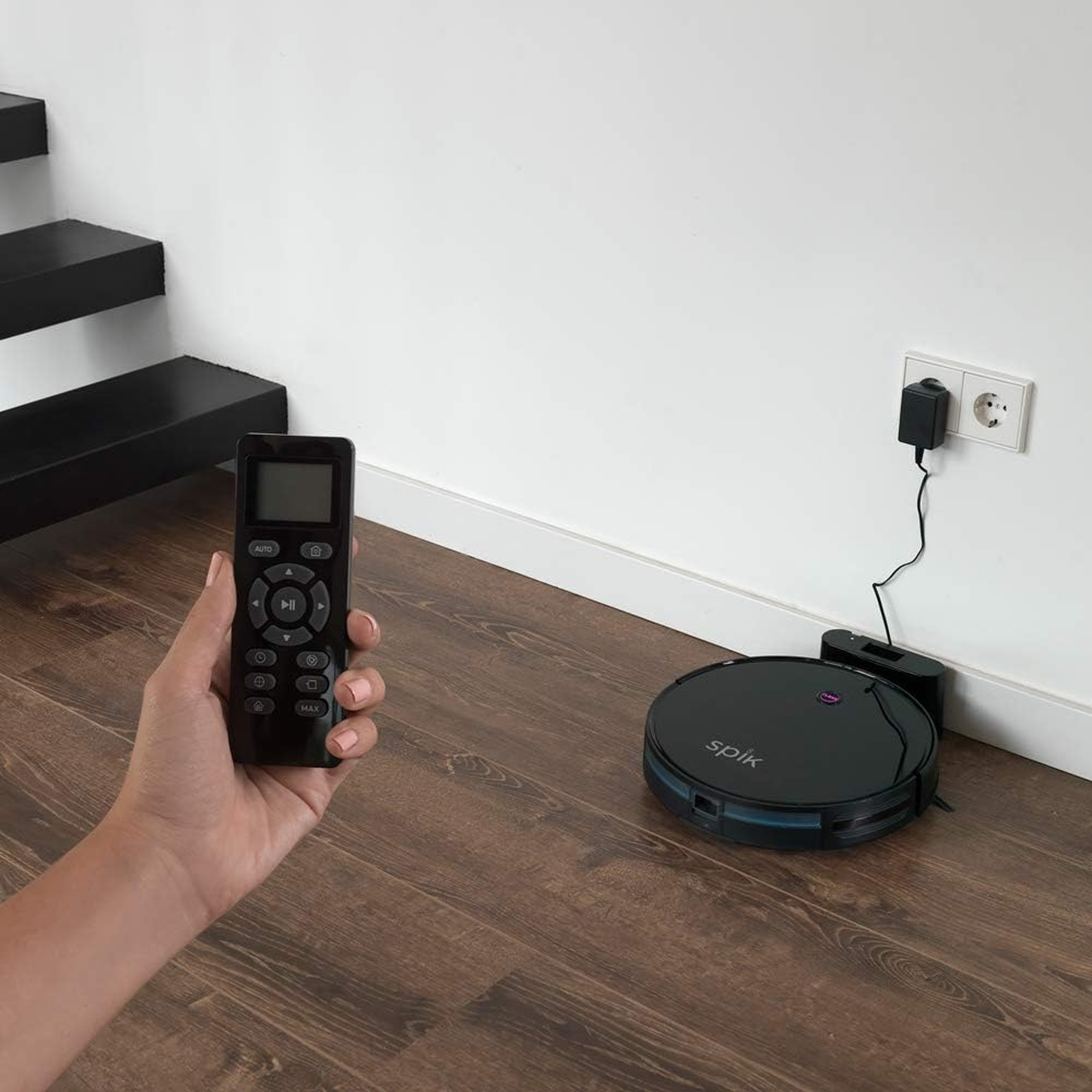 Una aspiradora robot, cargando, y una persona sosteniendo el control remoto.
