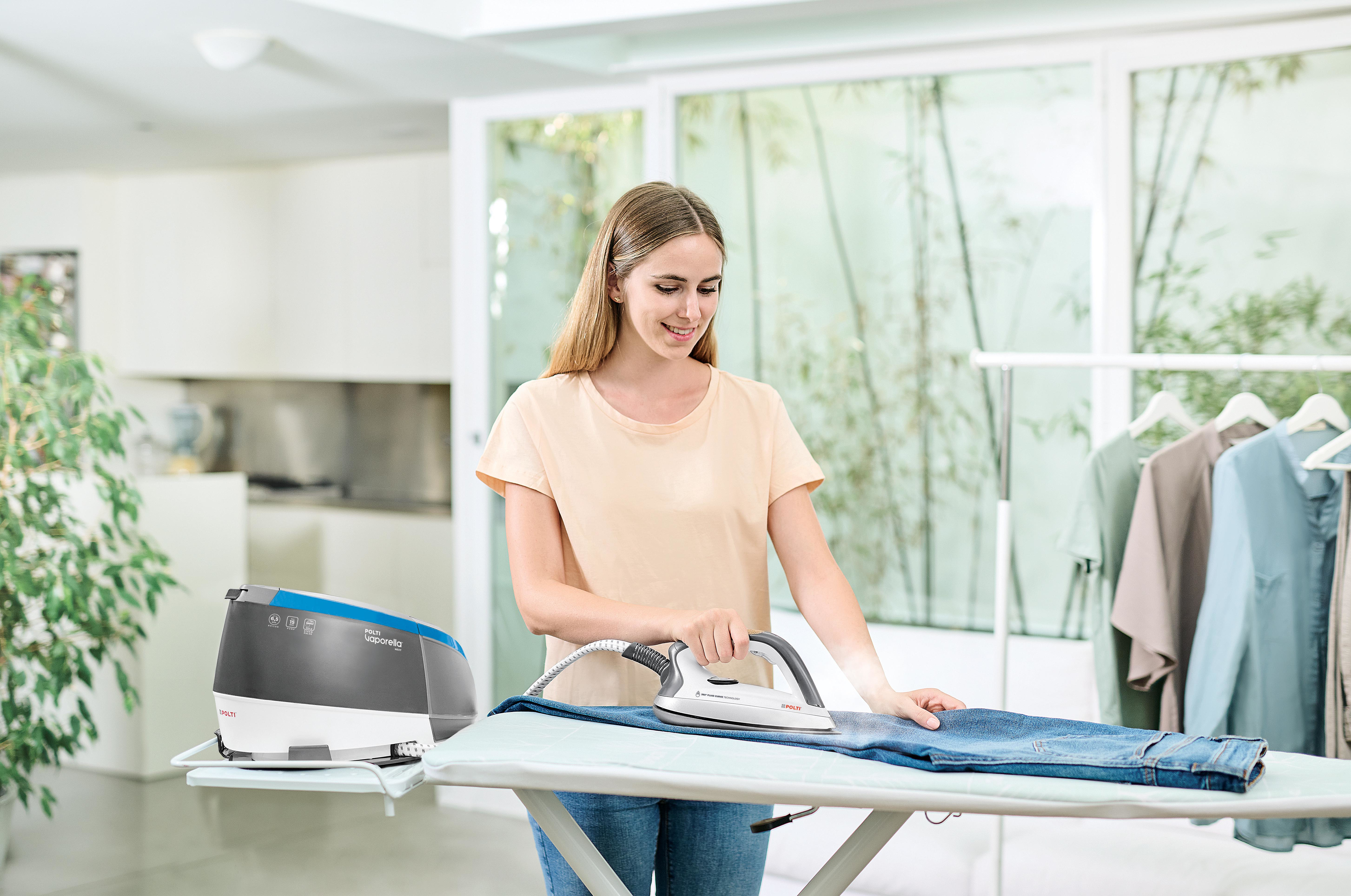 Donna stira jeans su asse. Ferro, lavatrice e vestiti appesi sullo sfondo.