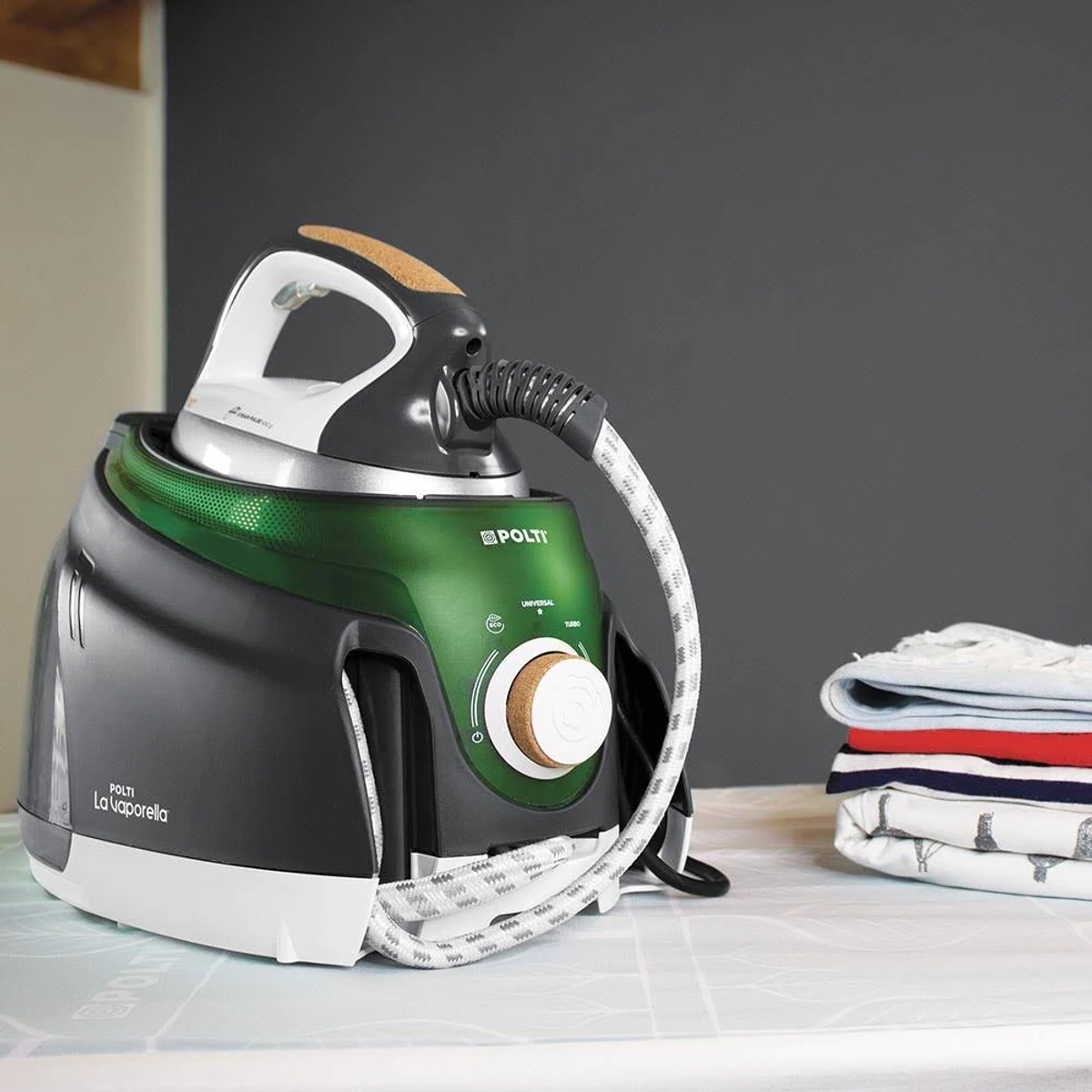 Un ferro da stiro nero e verde con cavo bianco, accanto a vestiti piegati su un tavolo.