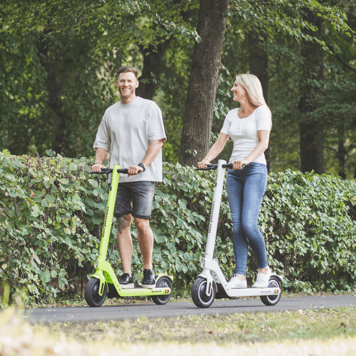 Zwei Personen fahren auf grünen und weißen Rollern auf einem gepflasterten Weg.