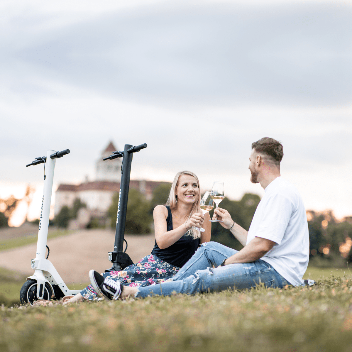 Zwei Personen stoßen mit Weingläsern an, die auf Gras sitzen, neben Rollern. Landschaft im Hintergrund.