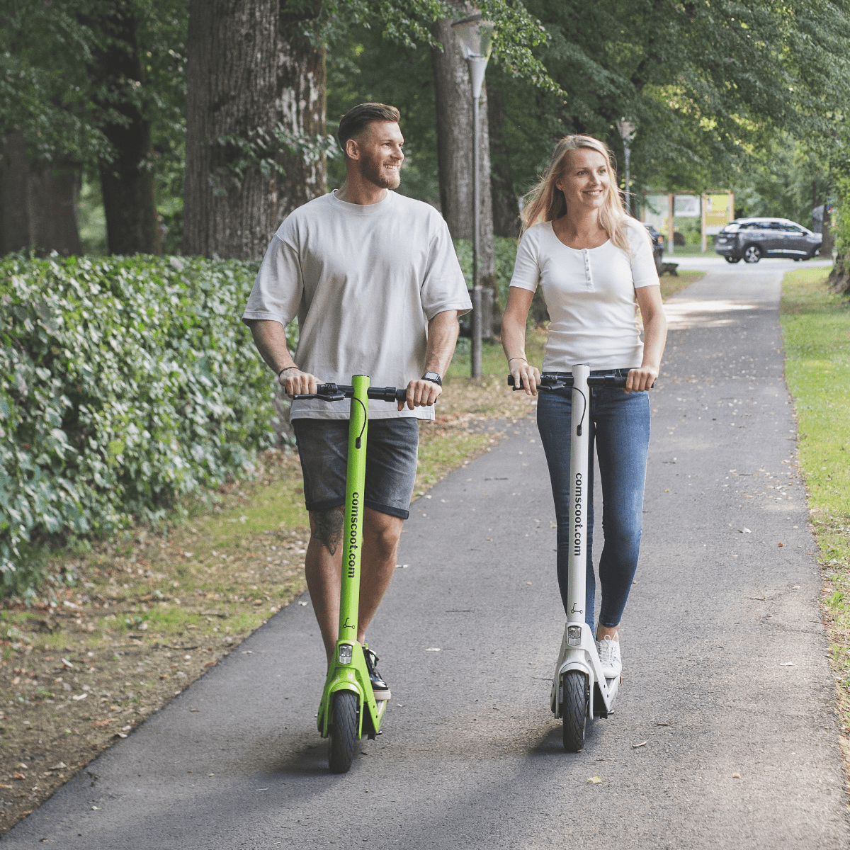 Mann und Frau fahren mit grünen und weißen Rollern auf einem Weg in einem Park mit Bäumen.