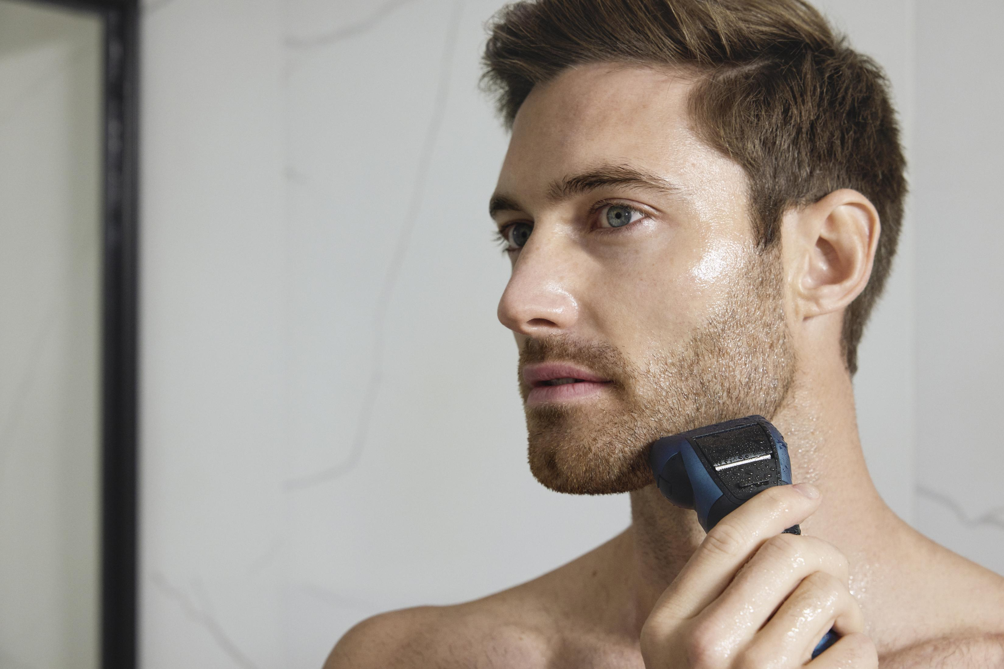 Mężczyzna goli się niebieską golarką elektryczną. Tło to rozmyta biała ściana.