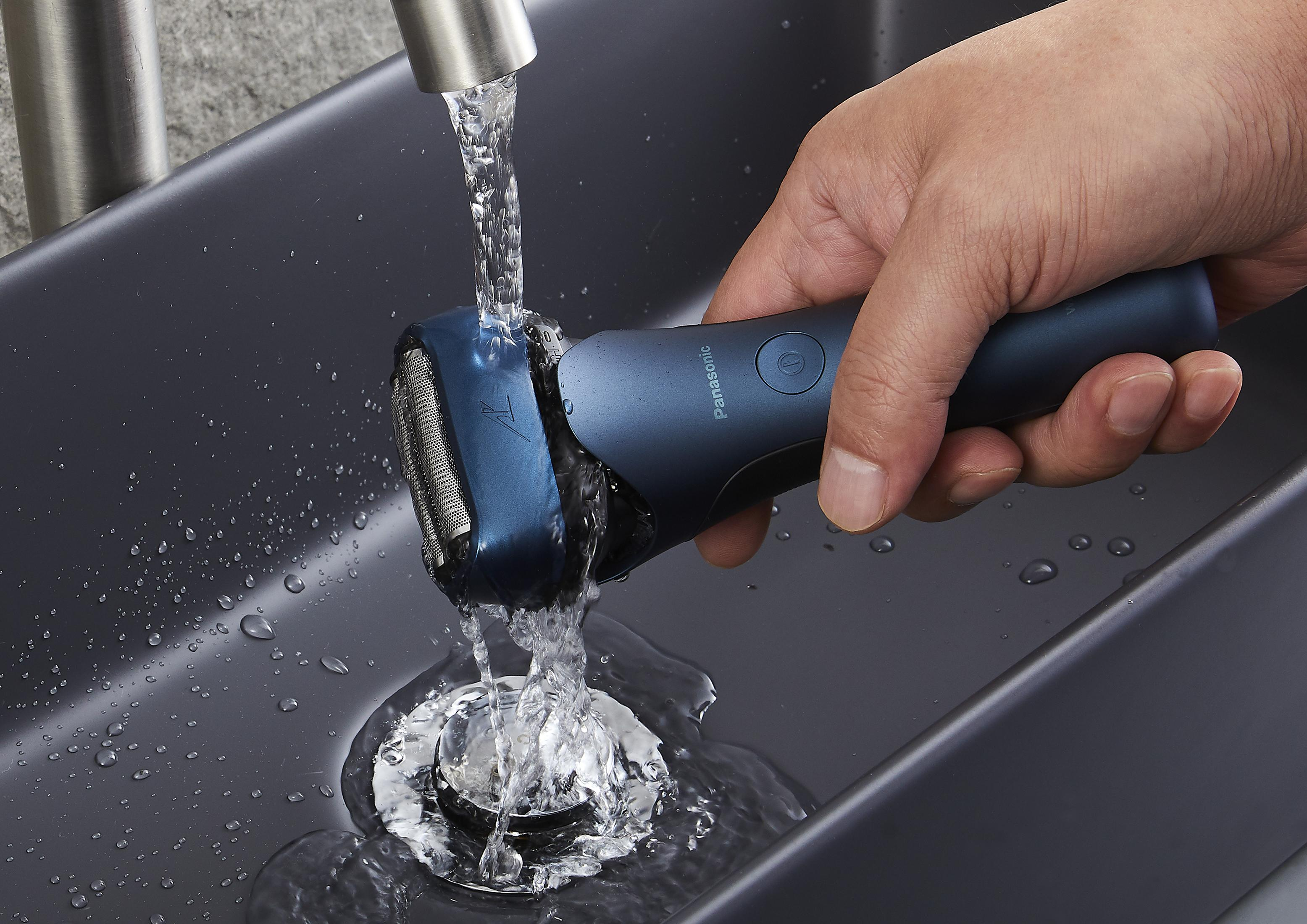 Dłoń płucząca niebiesko-czarną golarkę elektryczną pod bieżącą wodą w ciemnoszarej umywalce.