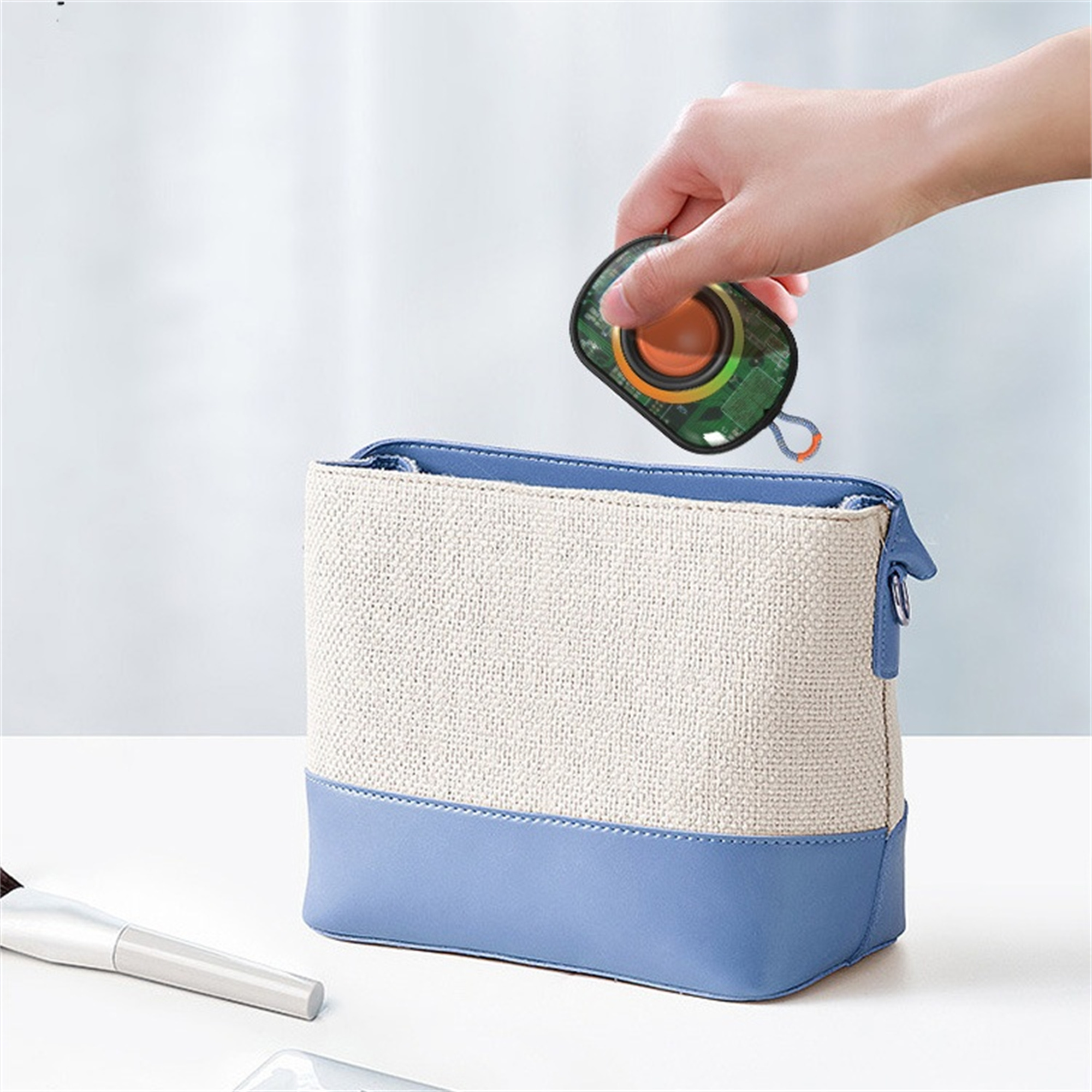 Eine Hand hält einen kleinen grünen Lautsprecher über einer blau-weißen Tasche. Es liegt auf einer weißen Oberfläche.
