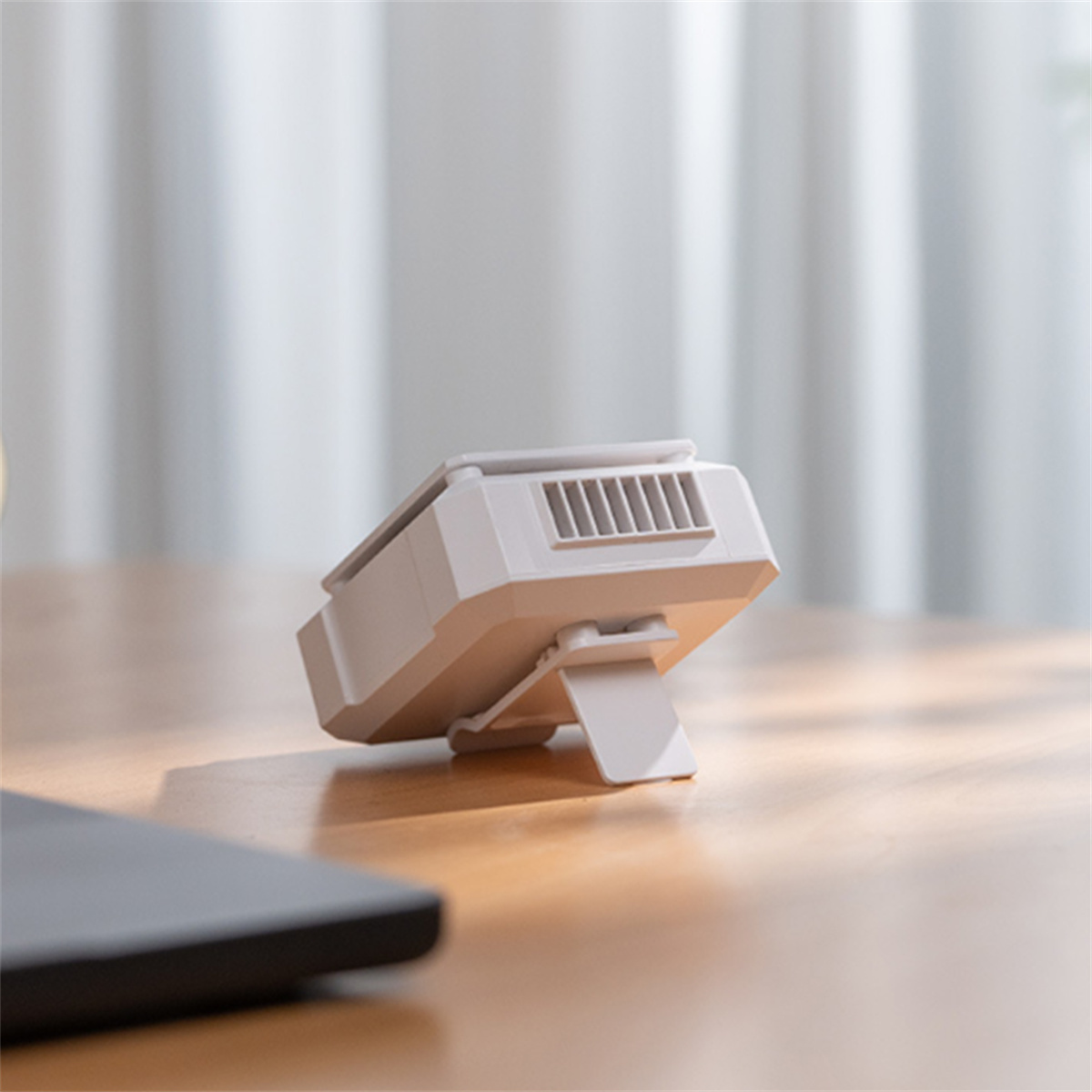 Ein kleiner, weißer Ventilator mit Ständer auf einem Holztisch, mit einem verschwommenen Laptop im Hintergrund.