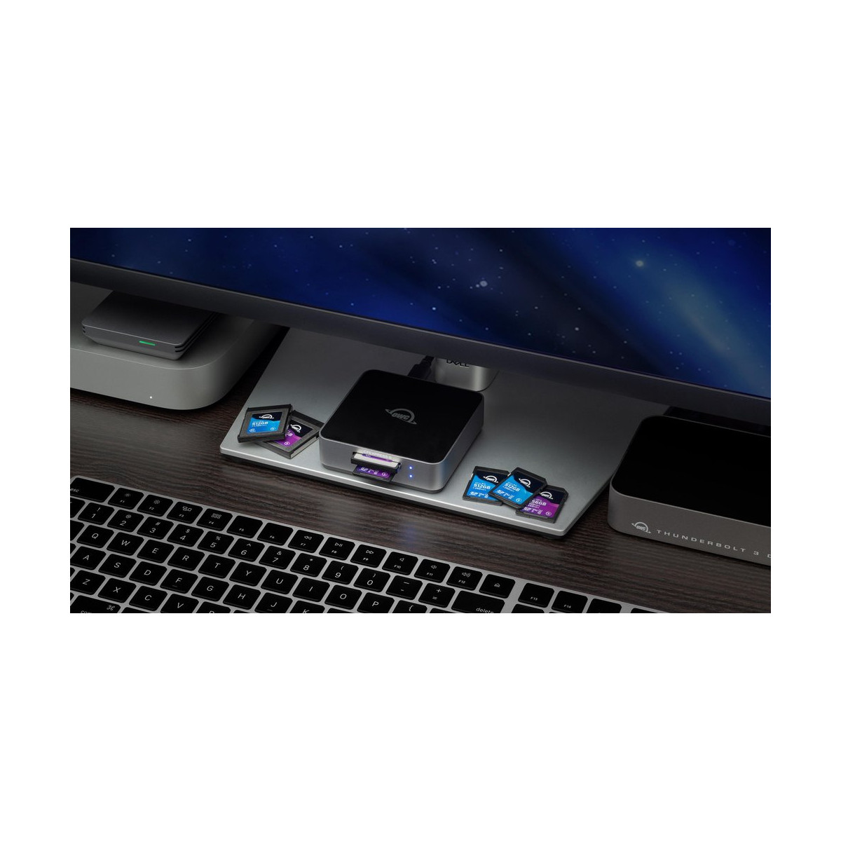 Konfiguracja biurka z monitorem, czarnym urządzeniem, kartami SD i klawiaturą. Ciemne drewniane biurko.