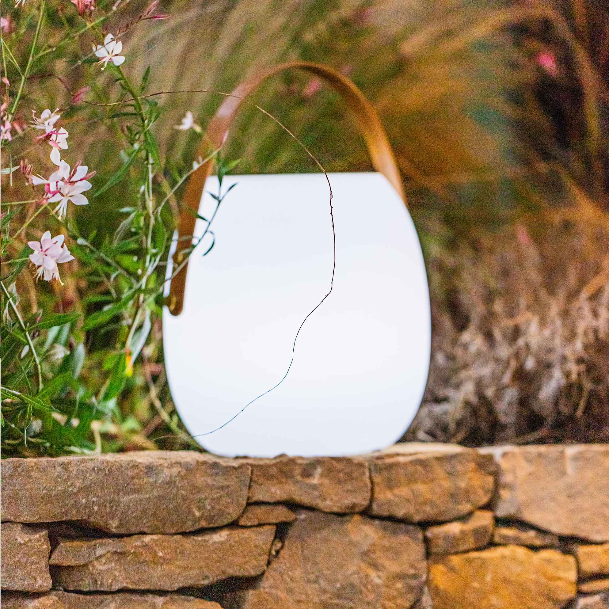 White lantern with a brown handle on a stone wall with flowers in the background.