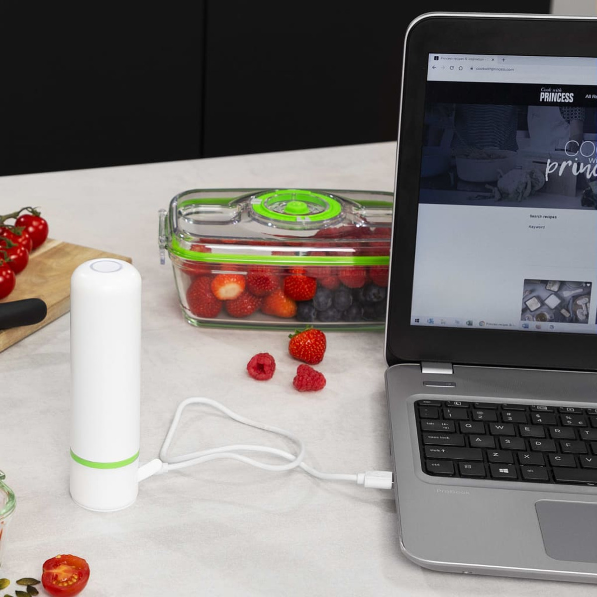 Ein Laptop, der mit einem Vakuumierer verbunden ist, mit einem Behälter mit Beeren und Tomaten.