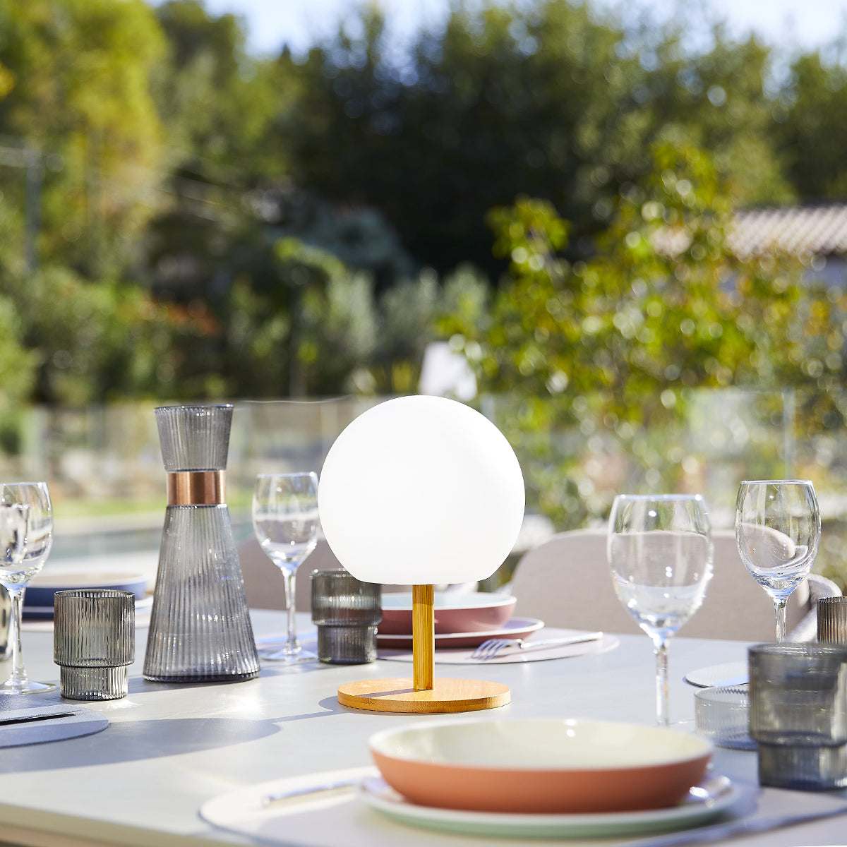 Ein Tisch für das Abendessen im Freien, mit einer weißen Kugellampe, Gläsern und Tellern an einem sonnigen Tag.