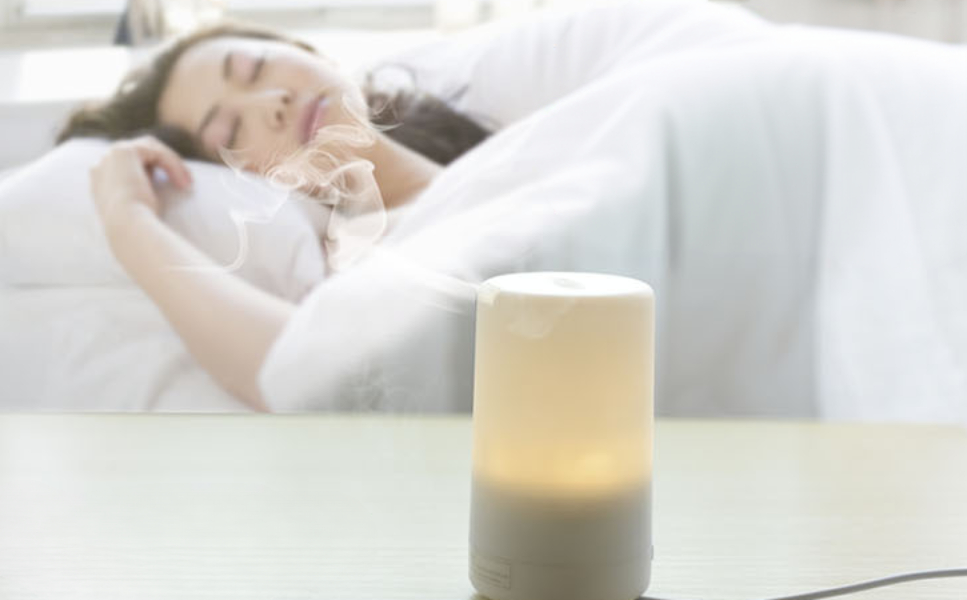 Aromatherapie-Diffusor auf dem Tisch, mit schlafender Frau im Bett im Hintergrund.