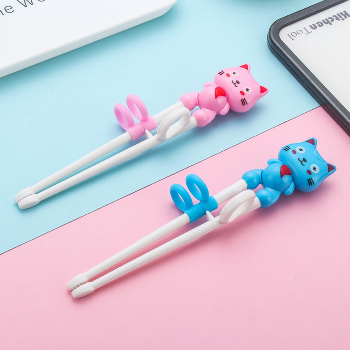 Zwei Essstäbchen aus Kunststoff mit Katzenfiguren in Rosa und Blau auf einer rosa und blauen Oberfläche.