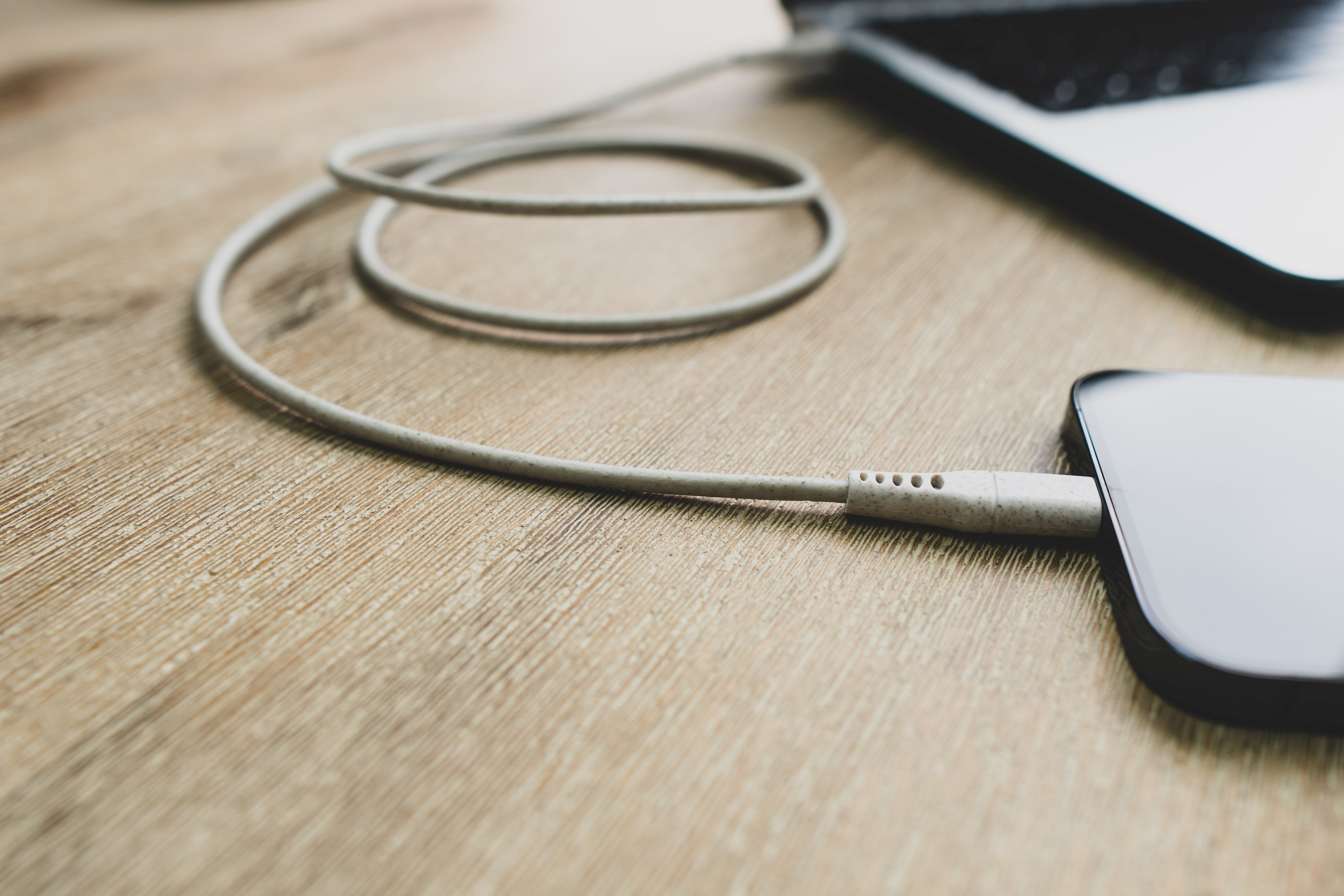 Telefoon opladen met een USB-C-kabel op een houten tafel, naast een laptop.