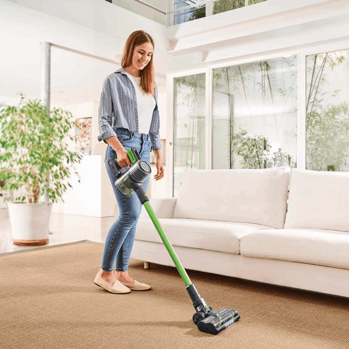 Mujer aspirando una alfombra con una aspiradora inalámbrica verde y negra. Sonríe.