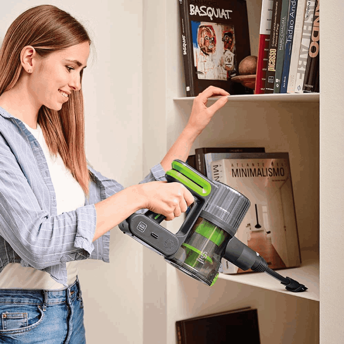 Mujer usando aspiradora, alcanza una estantería. Libros y estante con herramienta de limpieza. Estantería atrás.