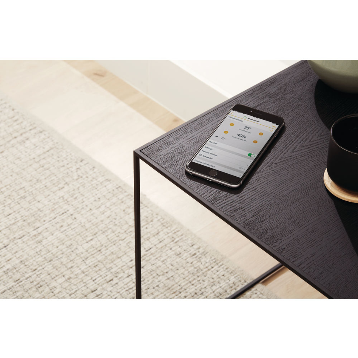 Un telefono su un tavolo nero mostra un'interfaccia smart home, accanto a un sottobicchiere e una tazza nera.