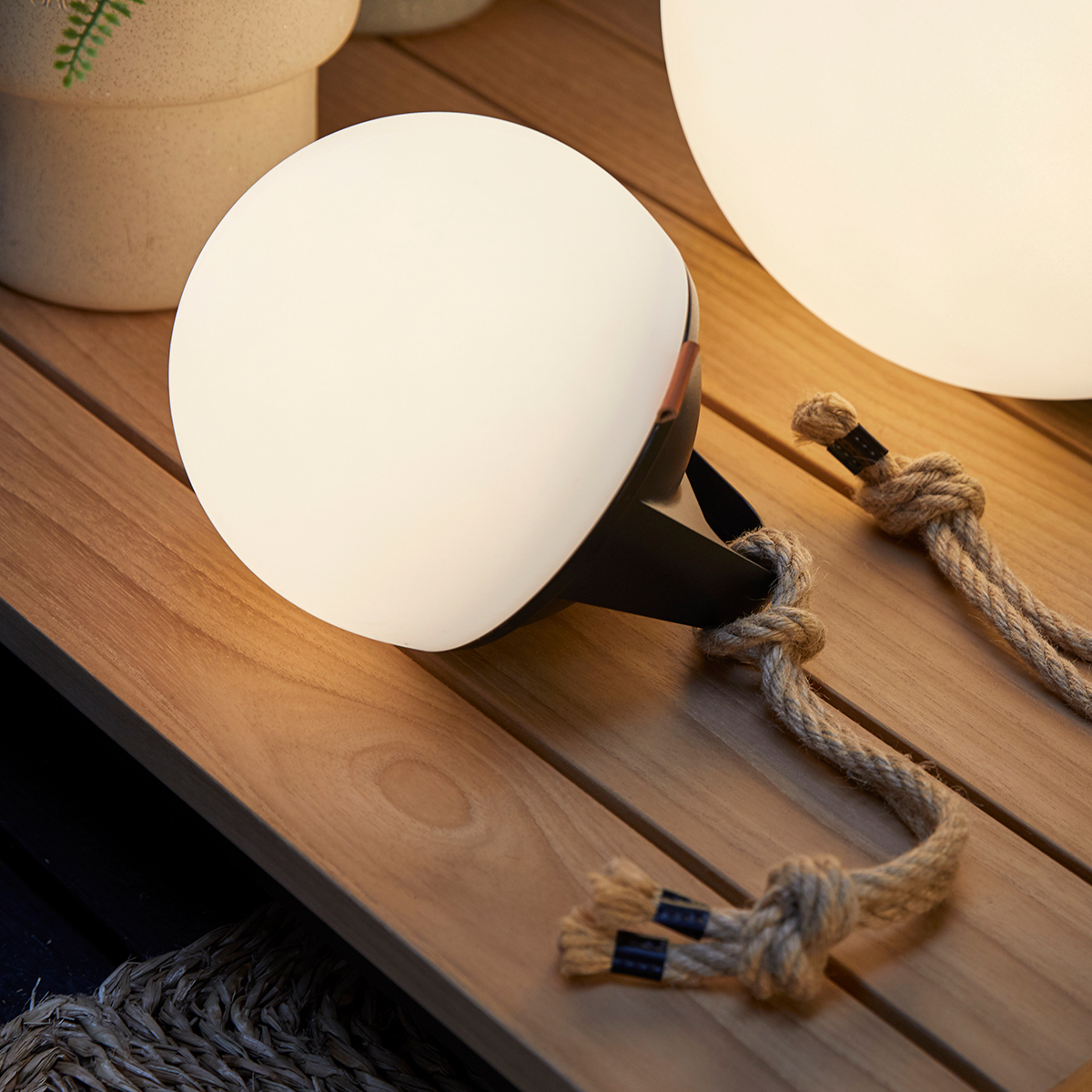 Leuchtende Kugel-Lampe mit schwarzem Sockel und Seildetails auf Holzoberfläche. Im Hintergrund eine weitere Kugel-Lampe.