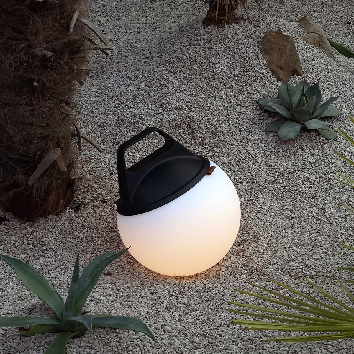 Eine weiße, runde Außenlampe mit schwarzem Griff auf einer Kiesoberfläche mit Pflanzen.