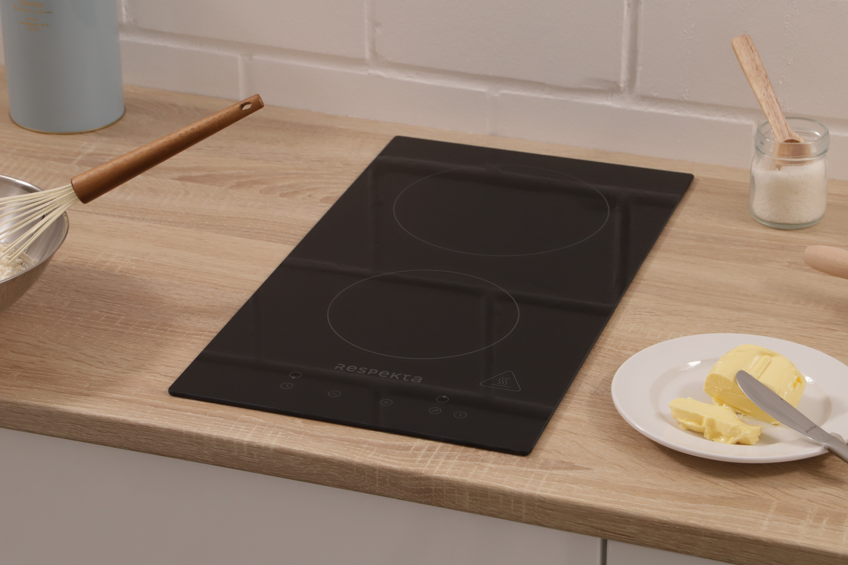 A black induction cooktop on a wooden surface. Butter and a knife are on a plate beside it.