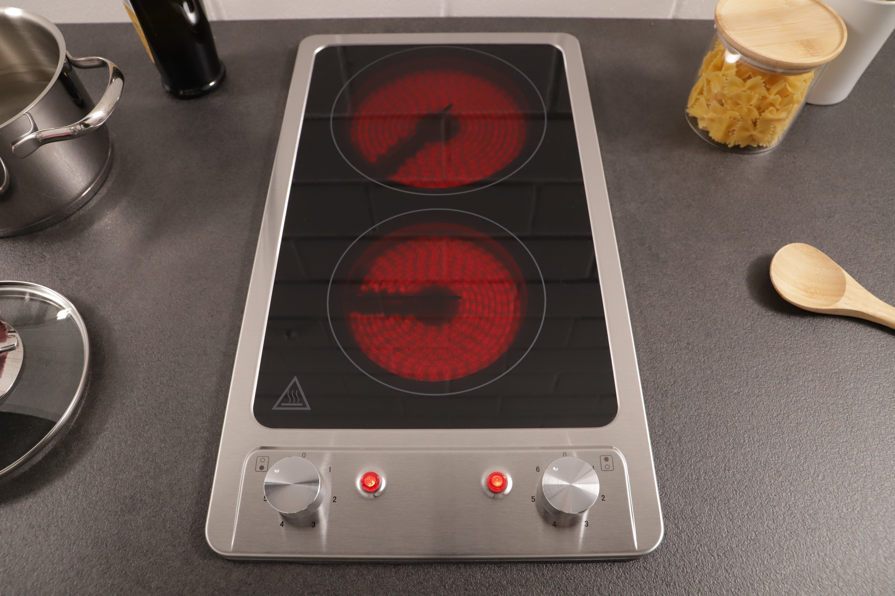 A silver electric cooktop with two red-glowing burners. Knobs and red lights are visible. A pot, spoon, and jar are in the background.