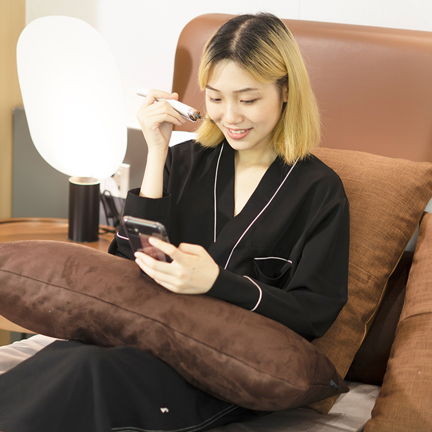 Frau benutzt ein Gesichtsgerät, während sie auf ihr Telefon schaut. Sie sitzt im Bett.